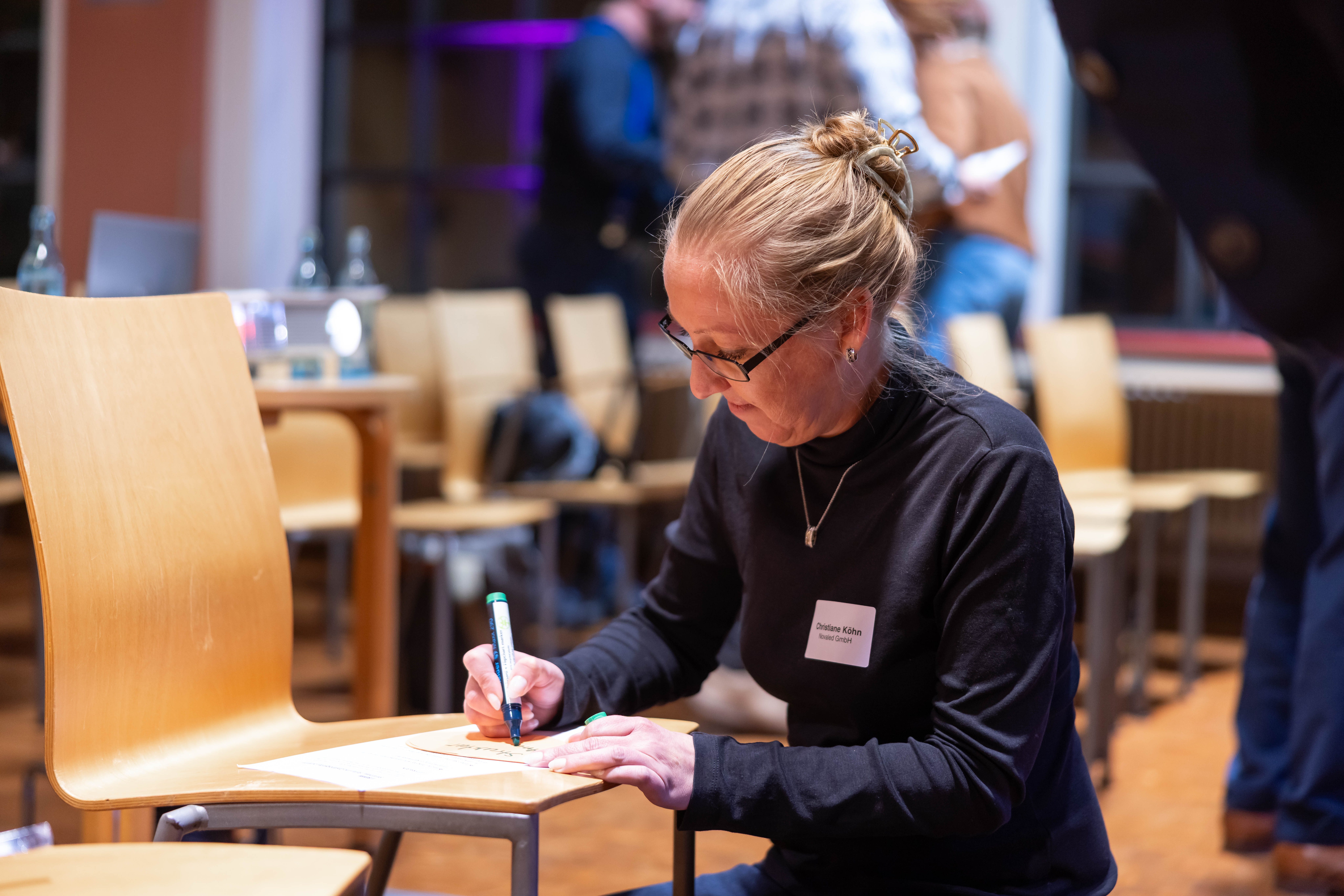 Eine Frau kniet vor einem Stuhl und schreibt auf der Sitzfläche auf einem Blattpapier.