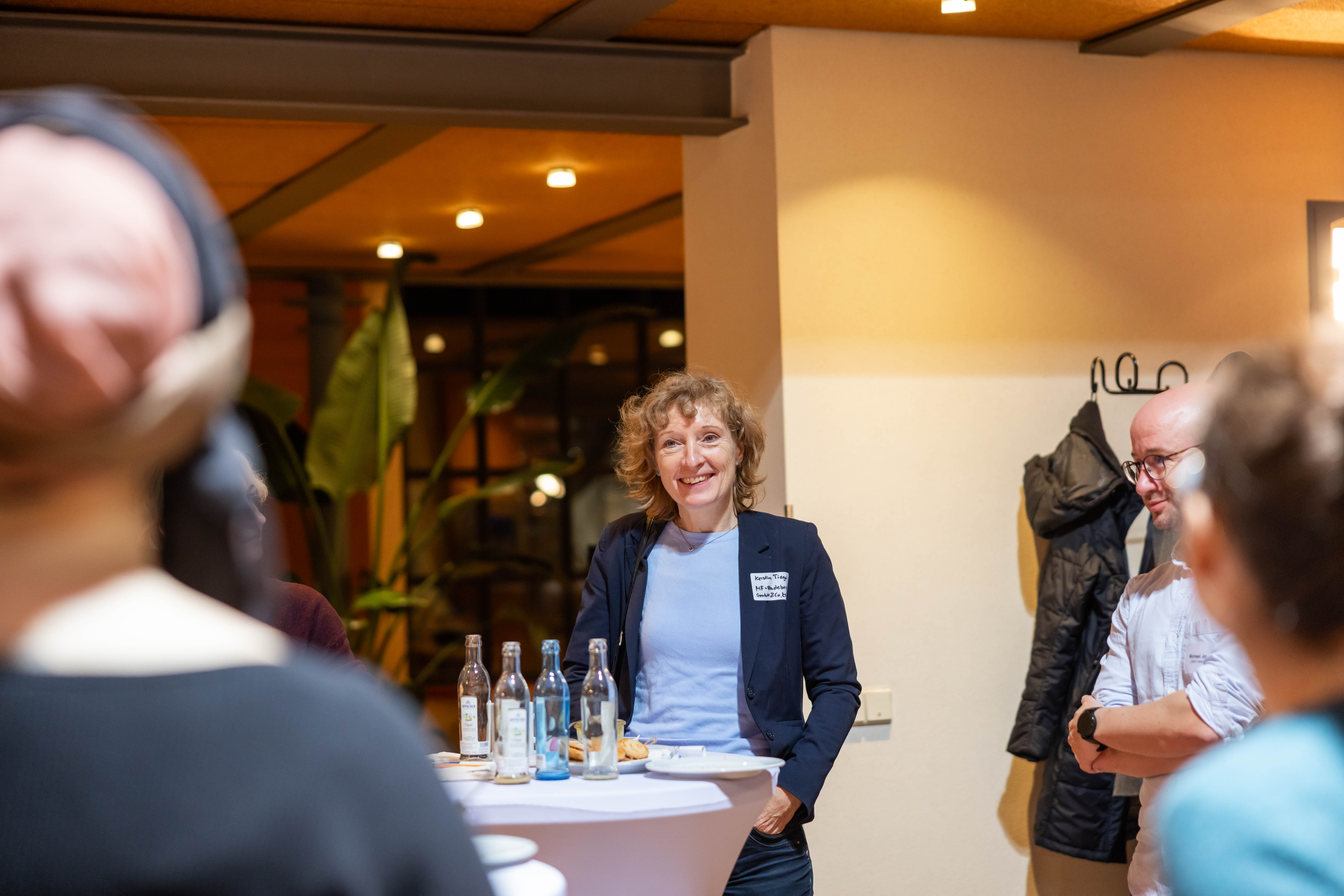 Eine Frau steht in einer Austauschrunde zusammen mit anderen Menschen und lächelt in die Kamera.